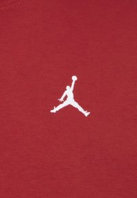 Tejido rojo con un logo Jumpman bordado en blanco. El diseño es suave con una textura sutil. En general, ofrece una estética deportiva.