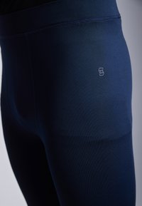 Sötétkék sport leggings sima textúrával, magas derékkal és egy kis logó részlettel a csípőn. Kényelem és rugalmasság érdekében tervezve.