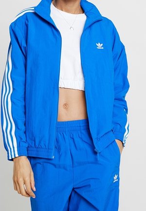 Personne portant une veste et un pantalon de survêtement Adidas bleus avec des bandes blanches et un t-shirt blanc court, main dans la poche.