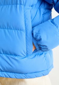 Vadderad blå jacka med en slät yta, med ficka fram och elastiska ärmslut. Synlig sömnad och en lätt glänsande finish.