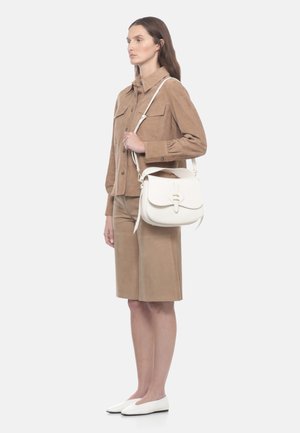 Donna con giacca beige e gonna al ginocchio tiene una borsa a tracolla bianca con dettaglio dorato, indossa scarpe bianche basse, rivolta verso sinistra.