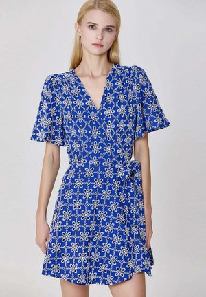 Femme blonde portant une robe portefeuille bleue avec broderies florales blanches et manches courtes volantées, debout devant un fond gris uni.