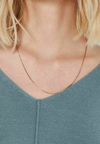 Una collana in oro riposa su un top azzurro chiaro a scollo a V, mettendo in mostra un design delicato e una texture liscia, con una leggera finitura lucida.