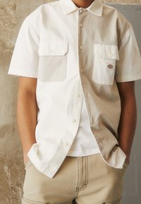 Camicia a maniche corte con colletto in cotone color crema, con due tasche sul petto e chiusura con bottoni. Indossata sopra una t-shirt bianca, abbinata a pantaloni beige.