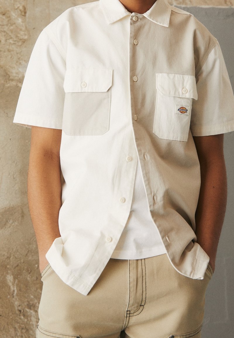 Camicia a maniche corte con colletto in cotone color crema, con due tasche sul petto e chiusura con bottoni. Indossata sopra una t-shirt bianca, abbinata a pantaloni beige.