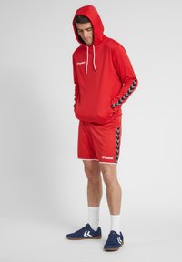 Rode sportieve hoodie met een frontzak en witte trekkoorden, gecombineerd met bijpassende rode shorts met zwarte chevronpatronen. Blauwe sportschoenen.