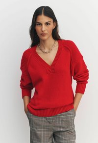 Red knitted sweater with a V-neck, ribbed cuffs, and hem. Features textured shoulder accents. Paired with checkered pants.