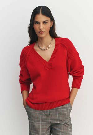 Red knitted sweater with a V-neck, ribbed cuffs, and hem. Features textured shoulder accents. Paired with checkered pants.