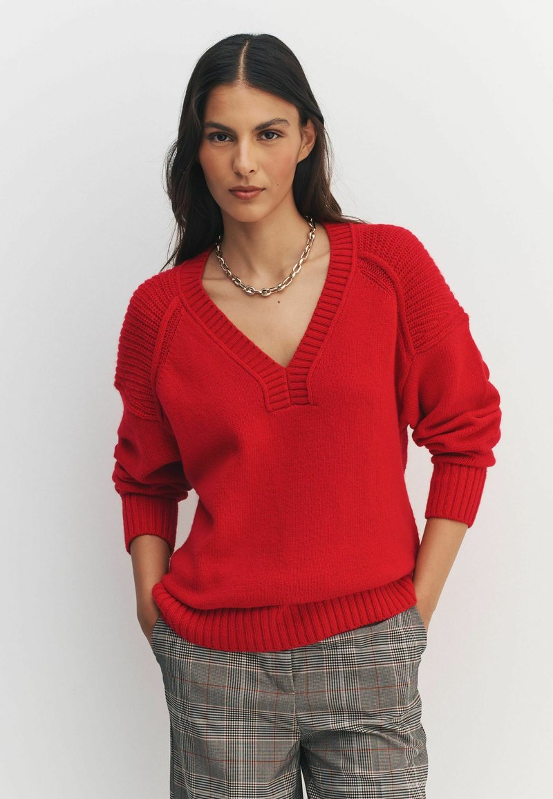 Red knitted sweater with a V-neck, ribbed cuffs, and hem. Features textured shoulder accents. Paired with checkered pants.