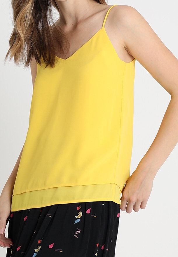 Femme portant un haut jaune sans manches à couches, associé à une jupe noire avec des motifs abstraits colorés.