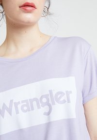 Levandulové tričko s krátkým rukávem, které má velké bílé obdélníkové logo s nápisem "Wrangler" tučnými fialovými písmeny. Příjemná měkká textura.