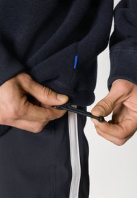 Mani che regolano il cordino elastico nero su una giacca in pile blu navy, indossata con pantaloni blu navy con strisce bianche sui lati, su uno sfondo neutro.