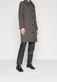 Persona con cappotto grigio scuro a quadri, pantaloni in pelle nera e stivali neri con tacco, in piedi su un pavimento grigio chiaro contro un muro bianco.