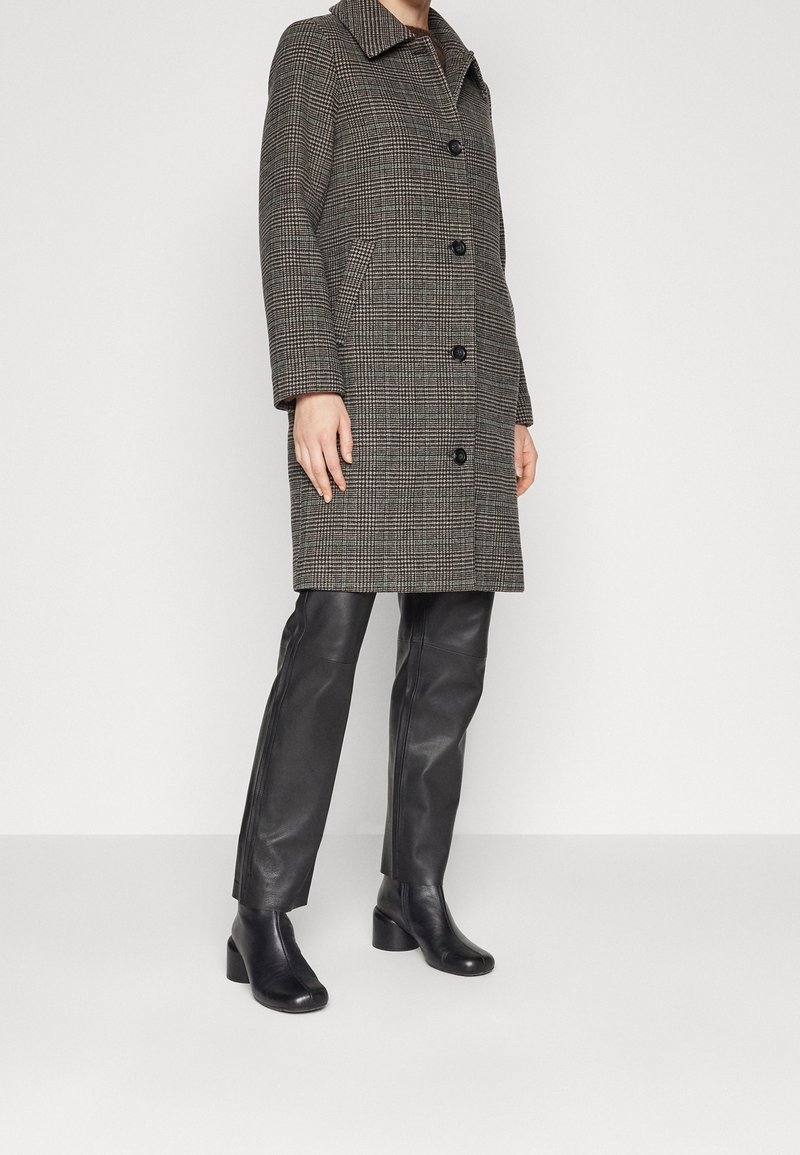 Persona con cappotto grigio scuro a quadri, pantaloni in pelle nera e stivali neri con tacco, in piedi su un pavimento grigio chiaro contro un muro bianco.