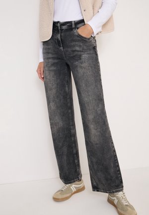 Persoon die versleten zwarte wijd uitlopende jeans draagt, beige sneakers, wit overhemd met lange mouwen en een beige fleecevest, hand in de zak, staand tegen een witte achtergrond.