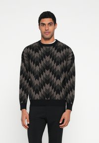 Brązowo-czarny dziergany sweter z zygzakowatym wzorem, okrągłym dekoltem, długimi rękawami oraz ściągaczami na mankietach i dole. W połączeniu z czarnymi spodniami.