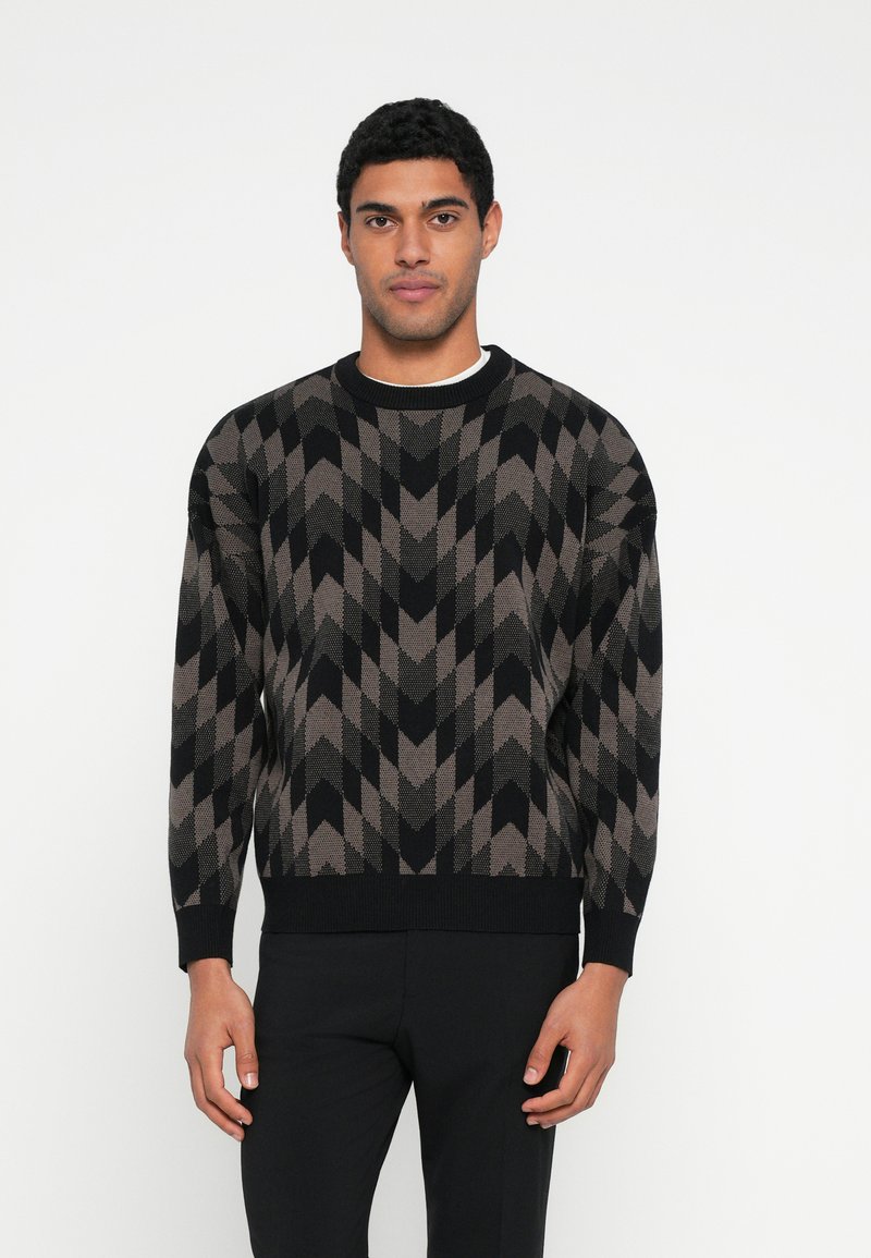 Brązowo-czarny dziergany sweter z zygzakowatym wzorem, okrągłym dekoltem, długimi rękawami oraz ściągaczami na mankietach i dole. W połączeniu z czarnymi spodniami.