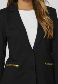 Blazer nero con chiusura a un bottone, rifiniture delle tasche in accento oro e design del rever classico. Indossato sopra una maglietta bianca.