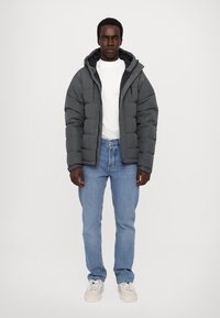 Doudoune grise avec capuche amovible, design matelassé, fermeture éclair, portée sur un t-shirt blanc et un jean bleu clair, associée à des baskets blanches.