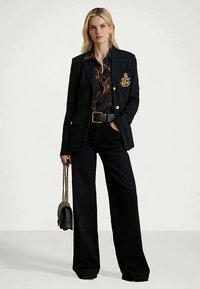 Mulher vestindo um blaser preto com brasão dourado, blusa preta com padrão, calças largas pretas, segurando uma bolsa preta com corrente dourada.