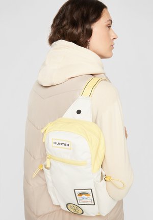 Kvinde med mørkt hår iført beige hættejakke og bærende en hvid og gul Hunter rygsæk over den ene skulder.