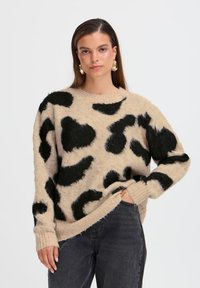 Pull en tricot beige avec un motif léopard noir duveteux, col rond et poignets côtelés, avec une coupe décontractée et un ourlet légèrement effiloché.