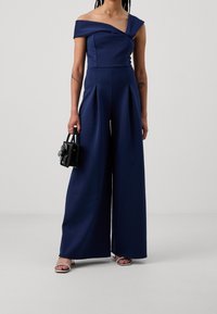 Jumpsuit azul marino sin hombros con piernas anchas plizadas, confeccionado en una tela suave. Combinado con un bolso negro que incluye un detalle de lazo. Tacones plateados.