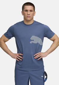 Blå atletisk t-shirt i et fugttransporterende materiale, har et stort hvidt logo af en springende kat på brystet, rund halsudskæring og korte ærmer.