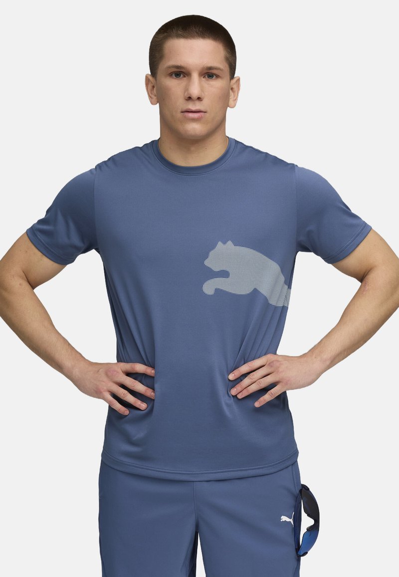 Blå atletisk t-shirt i et fugttransporterende materiale, har et stort hvidt logo af en springende kat på brystet, rund halsudskæring og korte ærmer.