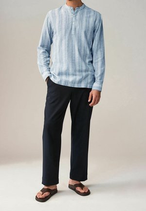 Mężczyzna ubrany w jasnoniebieską koszulę w paski z długim rękawem, czarne spodnie i brązowe sandały stoi z jedną ręką w kieszeni.