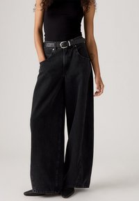 Zwarte wijde spijkerbroek van denim met voorzakken, een hoge taille, en gecombineerd met een zwarte riem. De stof heeft een lichte textuur.