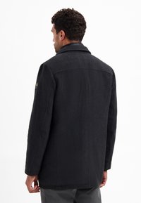 Zwarte wolmixjas met een reverskraag, gestructureerde schouders, rugnaden en een subtiele textuur. Kenmerkt zich door een logo badge op de mouw.