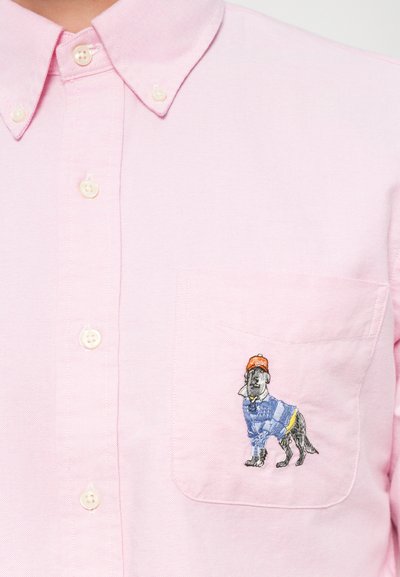 Chemise boutonnée rose clair avec une poche poitrine arborant un chien brodé portant une chemise bleue et une casquette orange. Présente un col arrondi et des boutons.