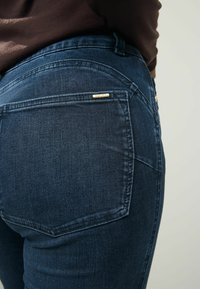Gros plan sur une personne portant un jean denim bleu foncé avec une poche arrière visible et un haut marron.