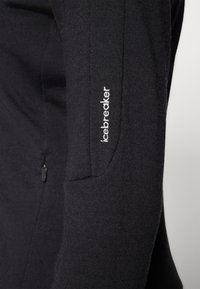 Melnas garām piedurknēm tops, kas izgatavots no mīksta auduma. Tam ir pieguļošs siluets, sānu rāvējslēdzēja kabata un balts izšūts "icebreaker" logo uz piedurknes.