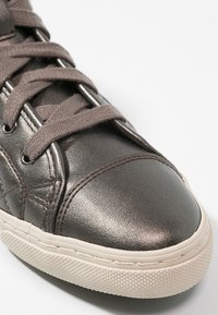 Metalické tmavě šedé tenisky s texturovaným koženým svrškem, kulatou špičkou a kontrastní béžovou gumovou podrážkou. Obsahují ploché tkaničky a prošívané akcenty.