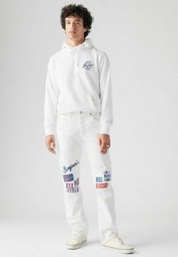 Witte hoodie met logo aan de voorkant, gecombineerd met witte denim broek met kleurrijke tekstprints. Texturen zichtbaar; casual stijl met sneakers.