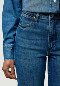 Primer plano de una persona que lleva jeans azul de cintura alta y una camisa de mezclilla metida, con una mano visible junto al muslo.