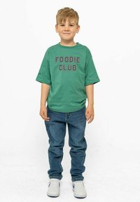 Zeleni pamučni majica s natpisom "FOODIE CLUB", u kombinaciji s plavim trapericama. Odjeća je opuštenog kroja i dolazi s bijelim tenisicama.