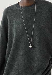 Pull en maille grise à coupe décontractée, arborant un long collier en chaîne argentée avec un pendentif métallique rond. Texture douce et design casual.
