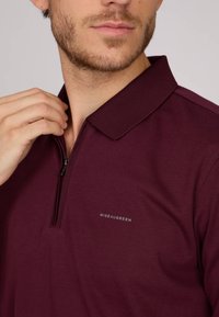 Polo burgundy en tissu lisse, avec un col zippé et un col texturé. Petit logo "Miseaugreen" sur la poitrine.