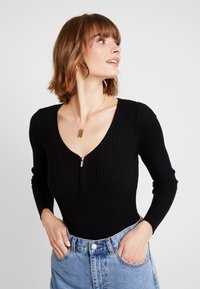 Zwarte geribbelde bodysuit met lange mouwen, diepe V-hals en een ritssluiting aan de voorkant; gecombineerd met een lichtblauwe denim rok met hoge taille.