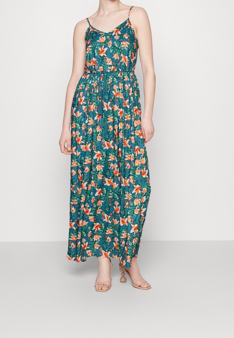 Kvinna klädd i en ärmlös teal maxi-klänning med orange blommönster och beige klacksandaler, stående mot en enfärgad grå bakgrund.