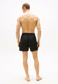 Pantaloni da nuoto neri con vita elastica, dotati di tasca posteriore e dettaglio logo discreto. Modello mostrato di spalle, a piedi nudi.