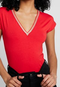 Roter Kurzarm-Body mit einem tiefen V-Ausschnitt und goldenen Akzenten. Der Stoff hat eine glatte Textur und wird mit hochtaillierten schwarzen Hosen kombiniert.