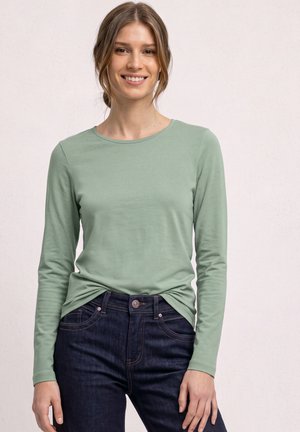 Jeune femme aux cheveux châtain clair en chignon bas, souriante, portant une chemise à manches longues vert clair et un jean bleu foncé sur un fond uni.