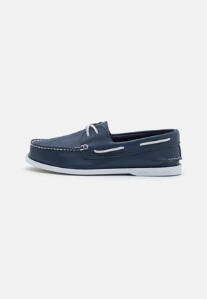 Chaussure bateau en cuir bleu marine avec lacets blancs, surpiqûres blanches et semelle en caoutchouc blanche sur fond uni.