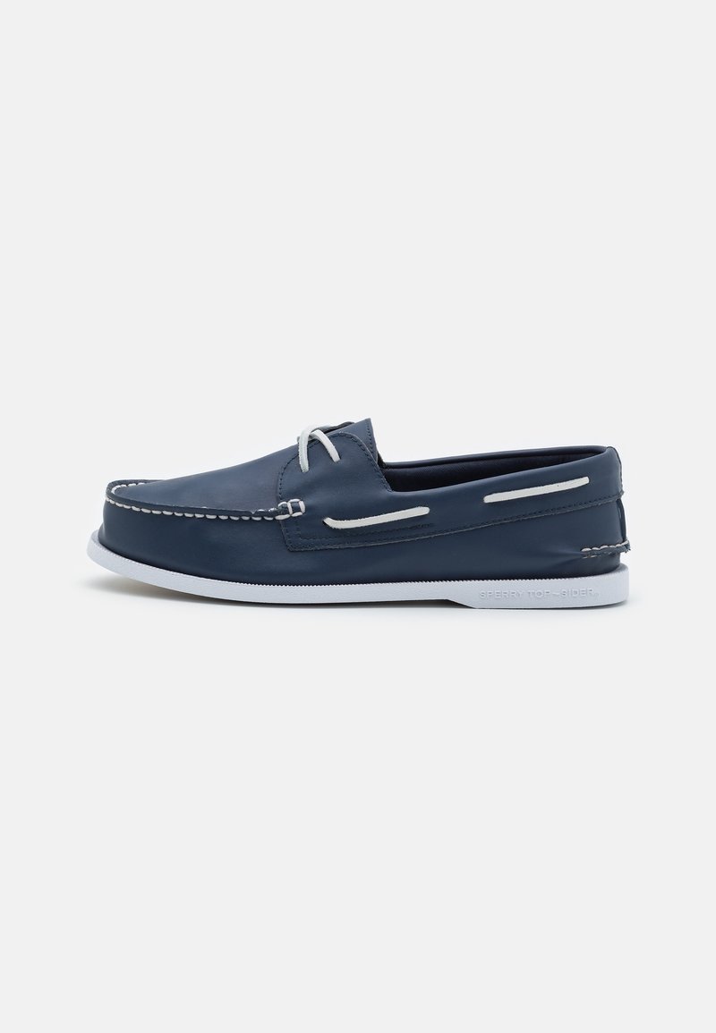 Chaussure bateau en cuir bleu marine avec lacets blancs, surpiqûres blanches et semelle en caoutchouc blanche sur fond uni.