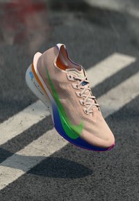 Zapatilla de running rosa claro con acentos verdes y azules, textura acanalada, suela acolchada y diseño de cordones planos. Bordes de suela de colores notables.