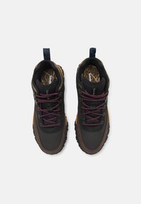 Chaussures de randonnée noires et marron avec des lacets violets, surfaces texturées et semelles robustes ; elles présentent une languette de traction bleue et des semelles intérieures à motifs.